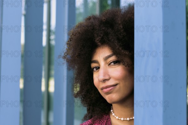 A young Arab woman with curly hair smiles confidently looking at camera, framed by blue columns. Her expression exudes warmth and charm, capturing a moment of poise and elegance