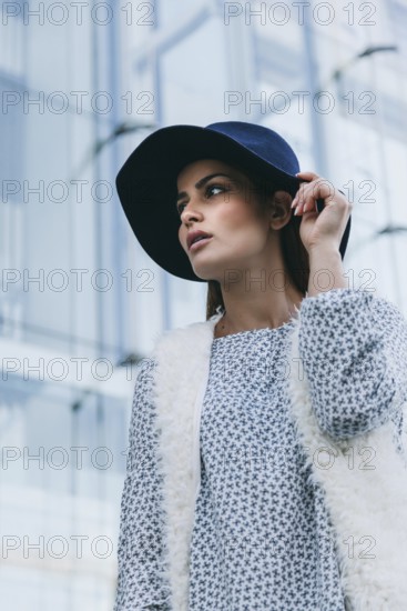 Elegant woman wearing a blue hat and fur vest, looking poised in an urban setting with glass buildings. Her stylish outfit features subtle patterns, exuding sophistication