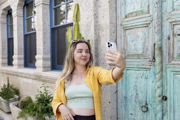 A woman takes a selfie on a charming street, wearing sunglasses and a yellow shirt. She stands in front of a rustic turquoise door, capturing the moment during a sunny day