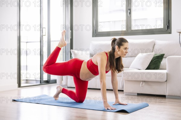 A pregnant woman in a red sports outfit practicing yoga on a blue mat indoors. She performs a pose focusing on wellness and relaxation, near a bright window and a comfortable couch