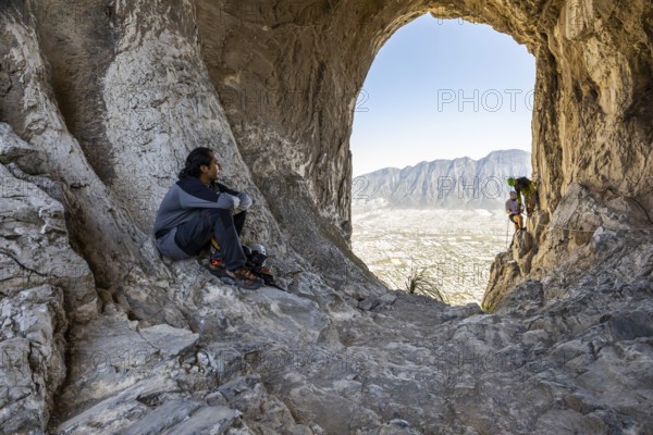 A climber rests on rocky terrain while two others practice mountaineering and rappelling in Eagleâ€™s Nest, Monterrey, Mexico. The cave opening frames the scenic mountains