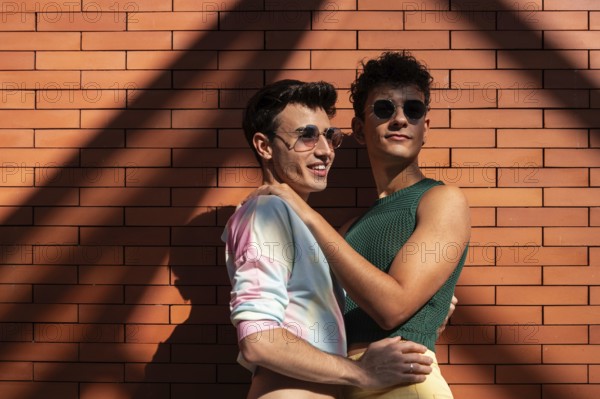 A happy gay couple in vibrant attire, embracing joyfully against a sunlit brick wall. They wear stylish sunglasses, capturing a moment of love and carefree intimacy