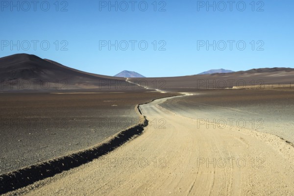 A desolate yet striking view of a dirt road meandering through the arid terrain of La Puna in Argentina, bordered by dark hills and a clear blue sky