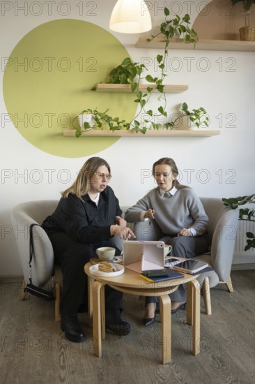 Two women engage in a business meeting at a cozy cafe. They are discussing something on a tablet, surrounded by coffee, cakes, and stylish decor with plants