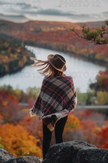A woman in a hat and plaid shawl stands overlooking a picturesque Quebec landscape in autumn. The vibrant red, orange, and yellow foliage create a stunning view
