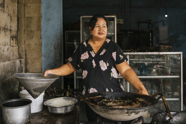 A woman stands in a rustic kitchen, preparing traditional dishes with a large wok. The worn walls and rustic tools create an authentic atmosphere, showcasing local cuisine of Bali