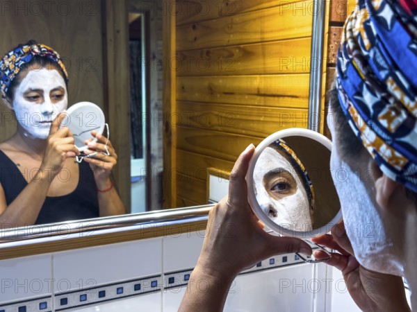A woman examines her skincare routine using a hand mirror in a cozy bathroom of Victoria, Australia. She wears a white face mask and colorful headscarf, emphasizing personal care and relaxation