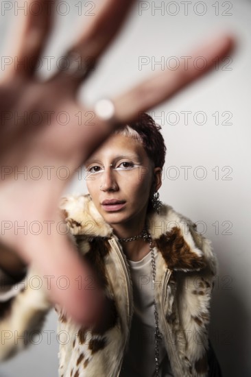 A woman wearing a stylish fur coat poses with hands stretched toward the camera. They sport bold makeup and multiple piercings, creating an edgy fashion statement