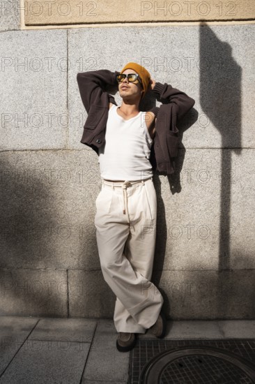 Stylish Colombian gay man wearing sunglasses and a beanie, leans against a city wall. His casual yet trendy outfit includes a tank top, cardigan, and wide-leg trousers