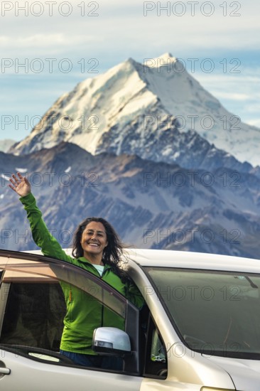 A woman stands joyfully in front of her car, surrounded by the breathtaking autumnal beauty of Mount Cook, New Zealand. The majestic mountain and vibrant colors enhance the scene