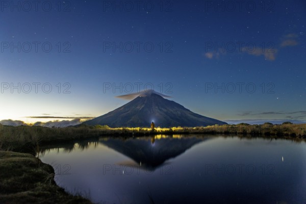 Back view of unrecognizable person gazing at Mount Taranaki's majestic peak reflected in a tranquil lake under a dawn sky, capturing New Zealand's serene natural beauty