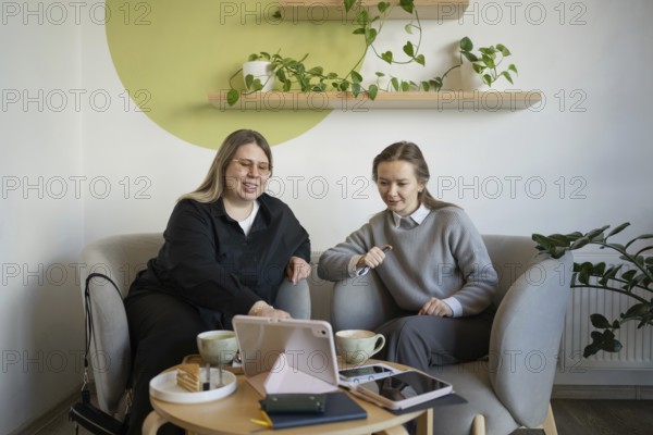 Two women sit in a cozy cafe, enjoying coffee and conversation. Laptops and notebooks are on the table, suggesting a mix of work and leisure in a relaxed environment