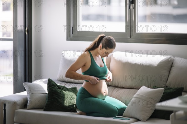 A pregnant woman, wearing a yoga outfit, sits on her knees on a couch, concentrating on well-being and mindfulness. She touches her belly