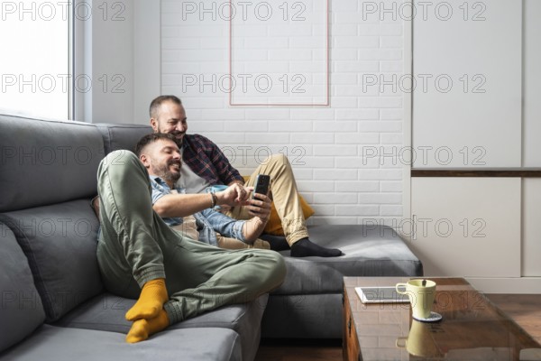 A happy gay couple relaxes on a cozy couch, smiling as they look at a smartphone. The setting is a modern, bright living room, exuding comfort and contentment