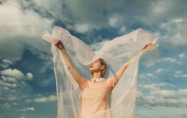 A woman with outstretched arms embraces the breeze, her sheer fabric flowing gracefully. A serene moment against a vivid blue sky, embodying freedom and joy