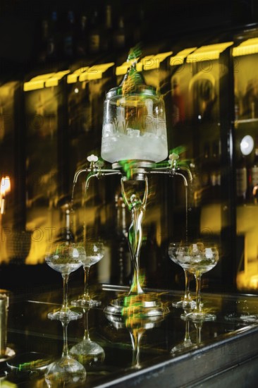 A vintage absinthe fountain pours into glasses on a dark bar counter. The ambiance is enhanced by a warm yellow glow, creating a classic and elegant atmosphere