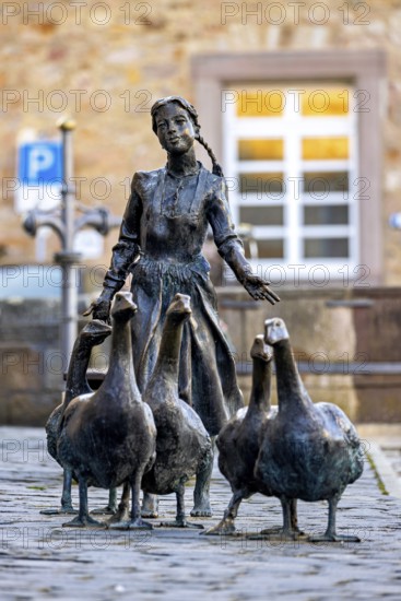 Bronze statue of a girl with geese on cobblestones, in the background a building with white windows, the Gänseliesel monument in Melsungen