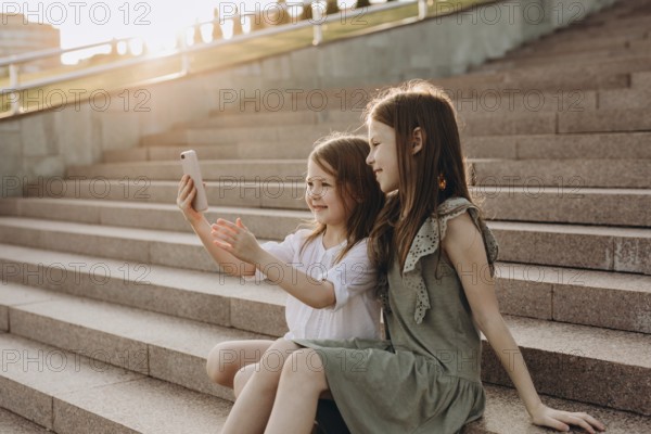 Two young girls sit on sunlit steps in summer, happily taking a selfie. They are enjoying a sunny day in a park, capturing moments of joy and togetherness with a smartphone