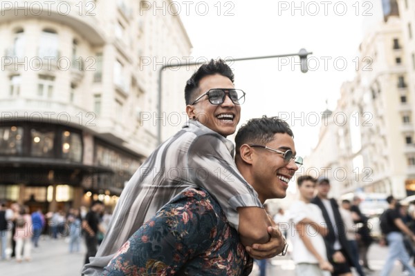 A cheerful latin gay couple enjoying a playful piggyback ride on a lively street in Madrid, surrounded by beautiful architecture and bustling city life