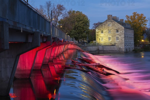 Illuminated Moulin Neuf water flow control dam and walkway over Des Mille-Iles river plus New Mill on Ile des Moulins in autumn at dusk, Old Terrebonne, Quebec, Canada