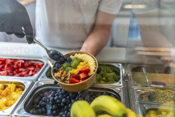Person wearing gloves preparing a nutritious acai smoothie bowl, adding fresh blueberries, sliced bananas, kiwi, mango, strawberries, and granola at a food counter