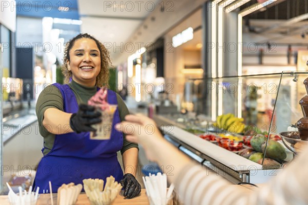 Young female entrepreneur serving a freshly prepared frozen yogurt cup to a customer, representing small business ownership and sweet service in a busy shopping mall environment