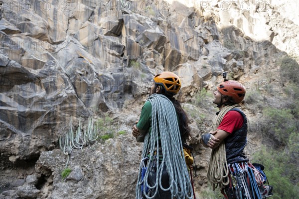Two climbers equipped with helmets and ropes study a rugged cliff, preparing for a traditional climbing adventure. The rocky landscape offers a challenging natural ascent