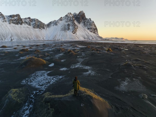 A man stands on a textured, frost-covered dune, gazing at the towering, snow-draped mountains of Stokksnes, Iceland, under a serene sunset sky
