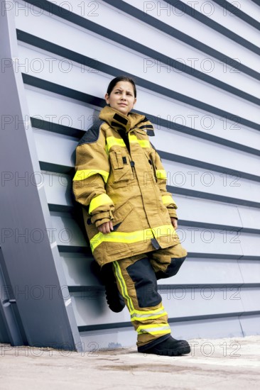 A young girl stands confidently against a modern wall while wearing a firefighter suit, showcasing strength and determination in an urban setting. Her expression is serious and focused