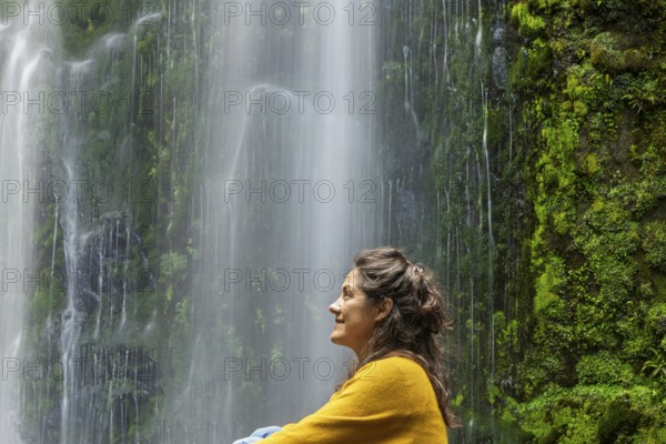 A woman in a yellow sweater enjoys a tranquil moment beside a lush green waterfall, surrounded by moss covered rocks. The scene evokes relaxation and nature beauty in the Great Ocean Road, Australia