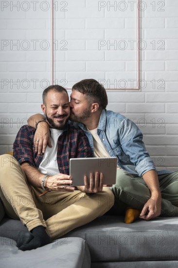 A loving gay couple sits on the couch, sharing a sweet moment while using a tablet. They embrace affectionately in a relaxed home setting, showcasing happiness and togetherness