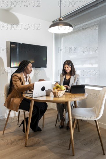 Two businesswomen are working together on their laptops and tablets in a hotel room, enjoying a healthy breakfast during their business trip