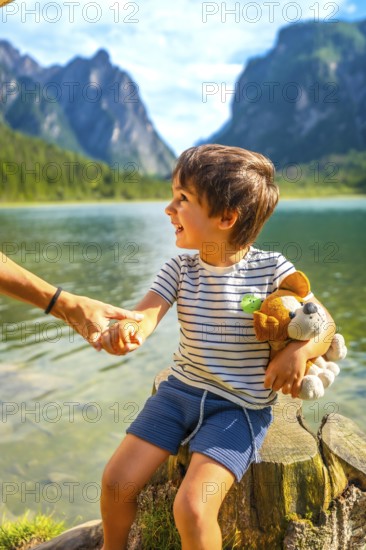 Smiling child holding a plush toy and his parent's hand while enjoying a summer day at lake dobbiaco with the dolomites in the background