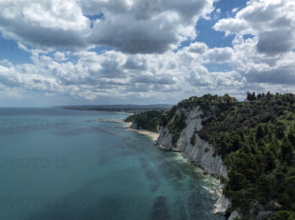 Aerial view of the stunning coastline in Sirolo, Italy. Dramatic cliffs rise from the turquoise sea, with lush greenery under a partly cloudy sky