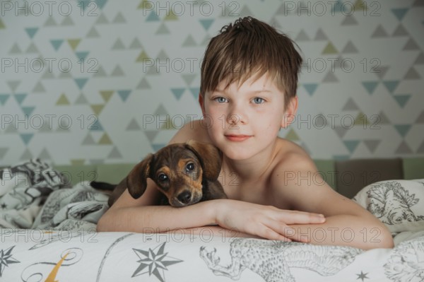 A young boy, with thoughtful blue eyes, gently holds his small Dachshund puppy as they relax together on a patterned bedspread. The intimate scene captures a tender moment of friendship and warmth in a softly colored bedroom