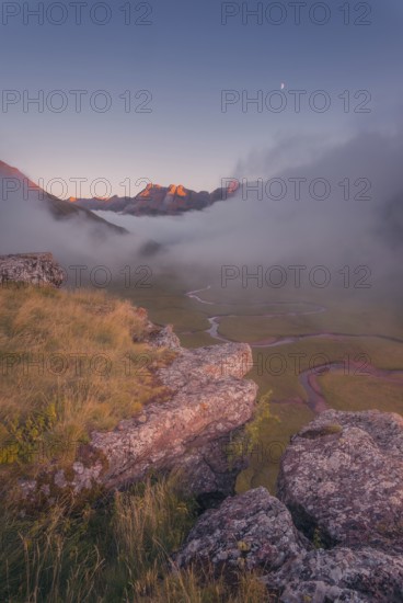 A breathtaking mountain landscape at dawn, with a rocky foreground, misty valley, winding stream, and glowing peaks in the Pyrenees. The moon hangs in the clear sky, adding tranquility