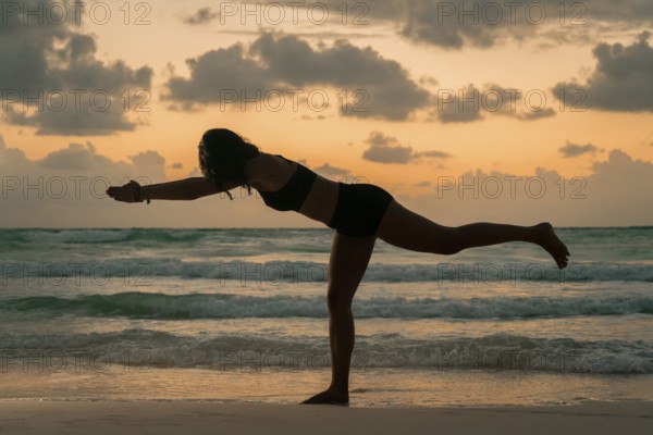A woman balances gracefully in a yoga pose on a beach, silhouetted against a stunning sunrise. The scene captures tranquility, focus, and the beauty of nature in Tulum, Mexico