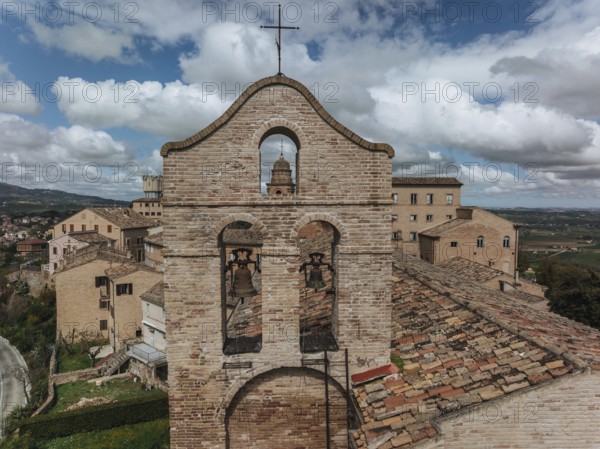 A historic bell tower stands amid rustic rooftops in Treia, Italy. The brick architecture contrasts with the lush countryside and blue skies in the background, ????, ????????, ??????