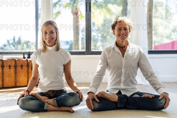 Male and female couple sits cross-legged on the floor in a sunlit room, meditating looking at camera. Both showcase relaxed expressions and a serene setting