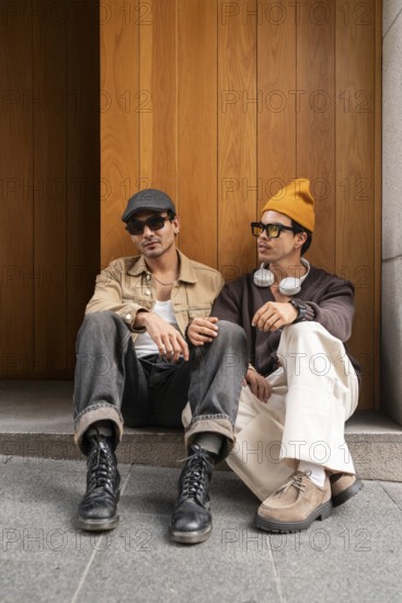 Two fashionable gay men sit on steps against a wooden backdrop. Both exhibit modern style with hats and sunglasses, emphasizing a relaxed urban vibe
