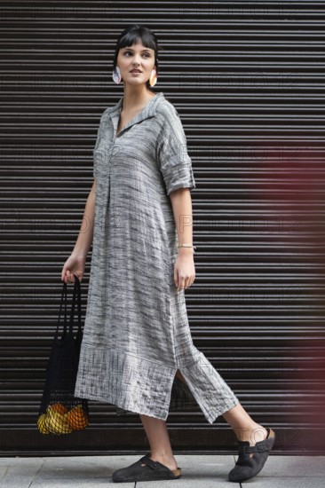 A fashionable woman walks confidently in the city while carrying a black bag with oranges and bananas. She is wearing a stylish gray dress and black shoes against a sleek metal background