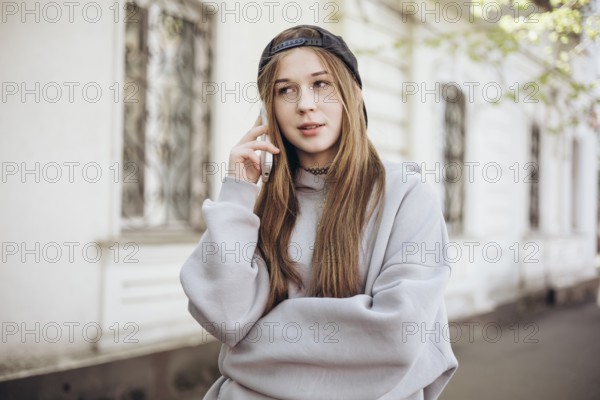A teenage girl wearing a casual hoodie and cap is engaged in a call on her smartphone outside. She appears relaxed and thoughtful, enjoying a moment in the open air