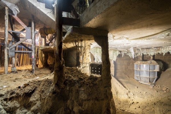 Illuminated underground construction site displaying active excavation work. The scene features supportive beams, exposed soil, and construction materials