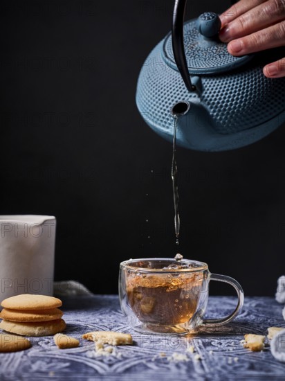 A serene setup of afternoon tea featuring a blue teapot pouring tea into a glass cup, accompanied by milk and a stack of cookies on a patterned tablecloth