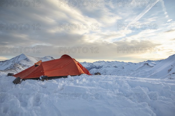 A vibrant orange tent sits atop a snow-covered peak, surrounded by majestic mountain views under a clear sky, ideal for ski touring or alpine adventure enthusiasts