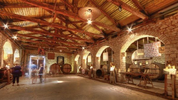 Interior view of a wine cellar with wooden roof and brick walls, few visitors, warmly lit, Achaia Clauss winery, Patras, Peloponnese, Greece
