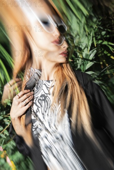 A stylish woman in sunglasses captured with motion blur effect, creating a dynamic and artistic look. She's surrounded by vibrant green leaves, adding to the visual interest