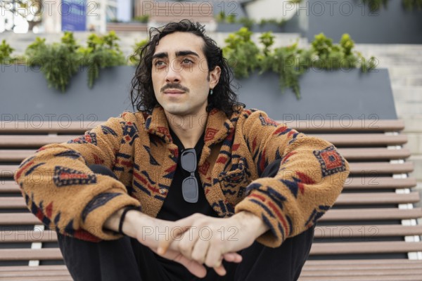 A man with curly hair and a mustache sits on an outdoor bench wearing a patterned jacket and sunglasses The urban background adds to his relaxed, stylish vibe