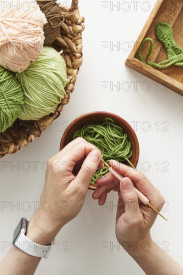 Top view of cropped unrecognizable hands crocheting with a pink hook and green yarn in a creative studio. A basket of yarn sits nearby, showcasing artisan needlework