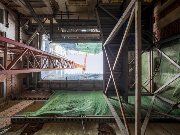 Looking up inside a building under construction, steel beams, scaffolding, and a towering crane frame the sky. Green safety netting covers areas for protection